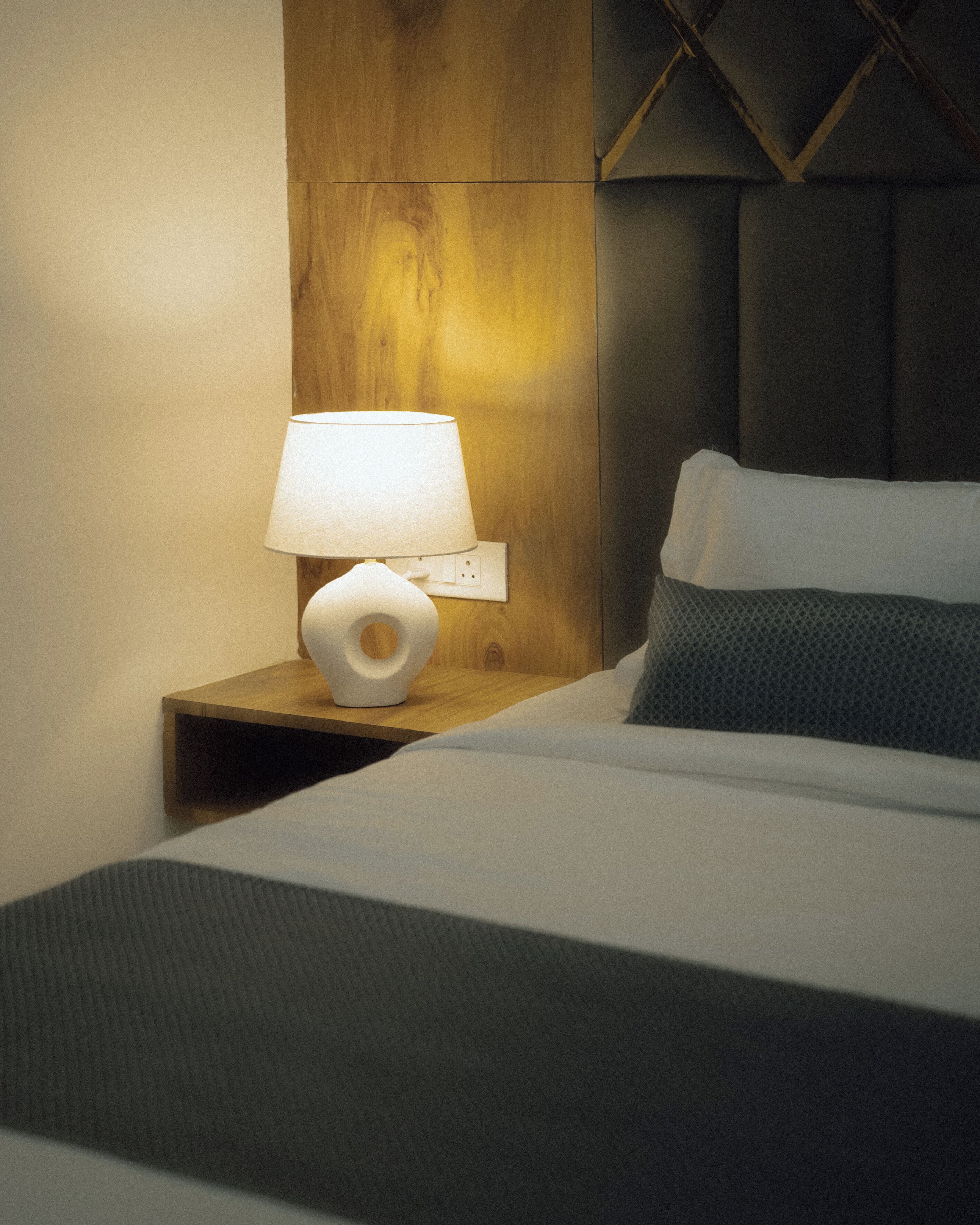 Een keurig opgemaakt bed met een wit-grijs dekbed, een houten nachtkastje met een witte lamp en een grijs hoofdbord.