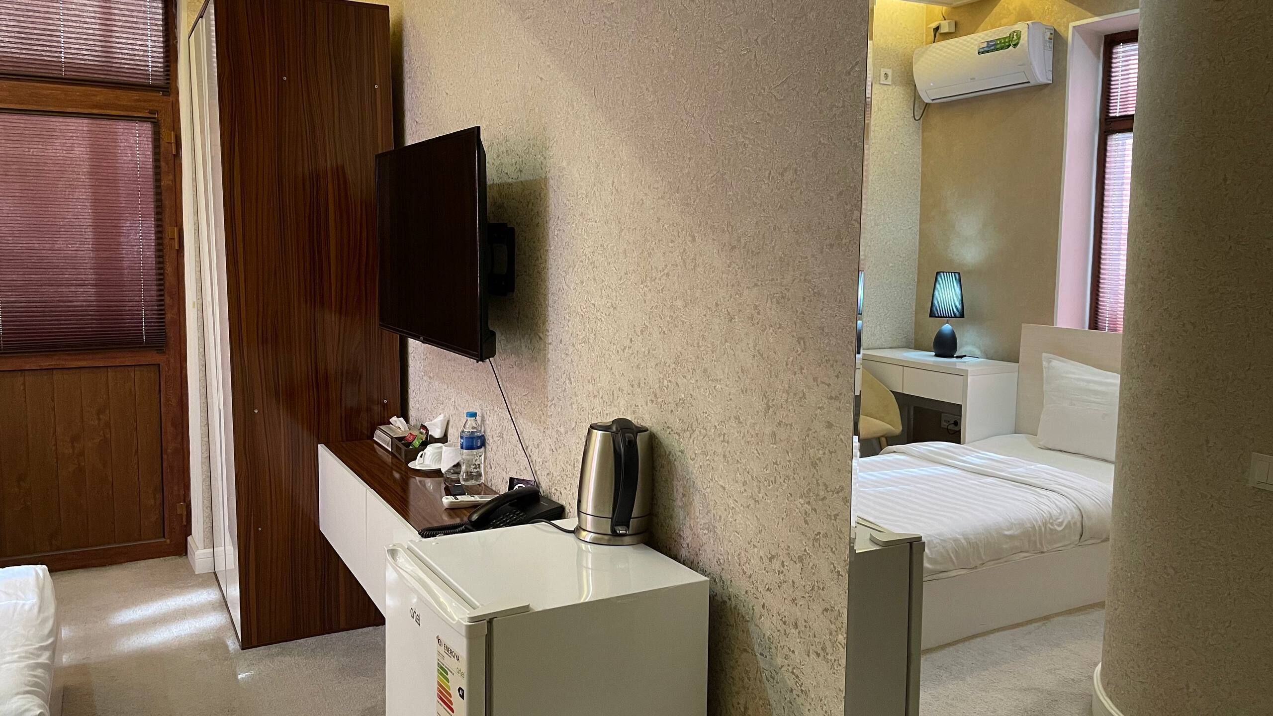 A hotel room with a wooden door, a desk with a flat-screen TV, a minibar, a bed, and a window with blinds.