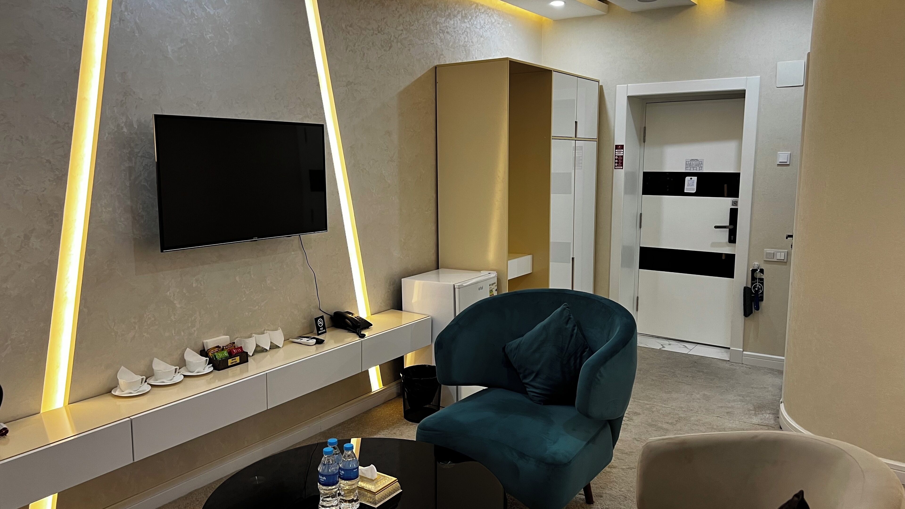 Hotelkamer met een flatscreen televisie aan de muur, een minibar, een zithoek met een blauwgroene fauteuil en een deur naar de badkamer.
