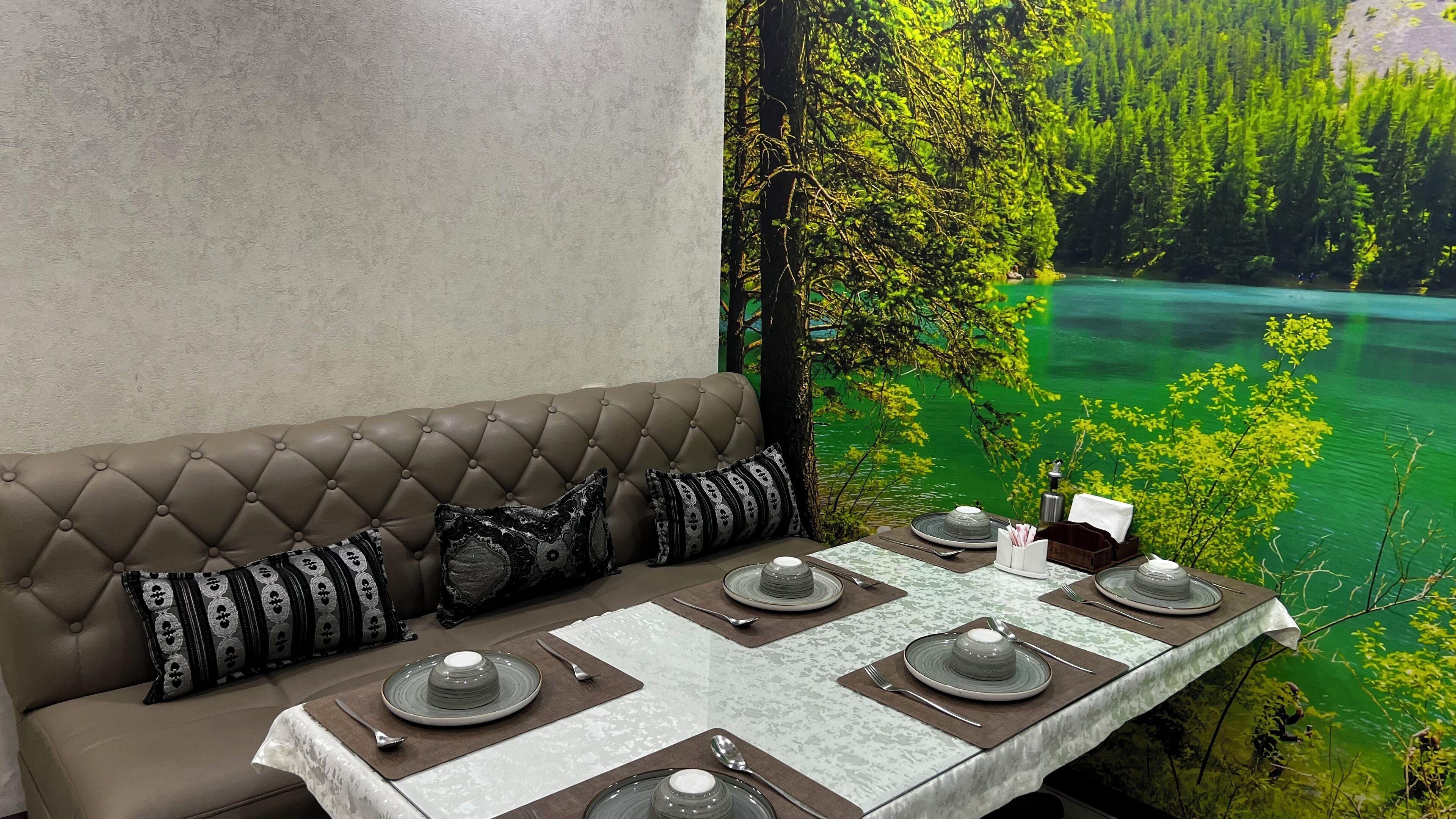 Ein Essbereich mit einem Tisch für vier Personen, einer Couch mit gemusterten Kissen und einem Wandbild von Wald und See.