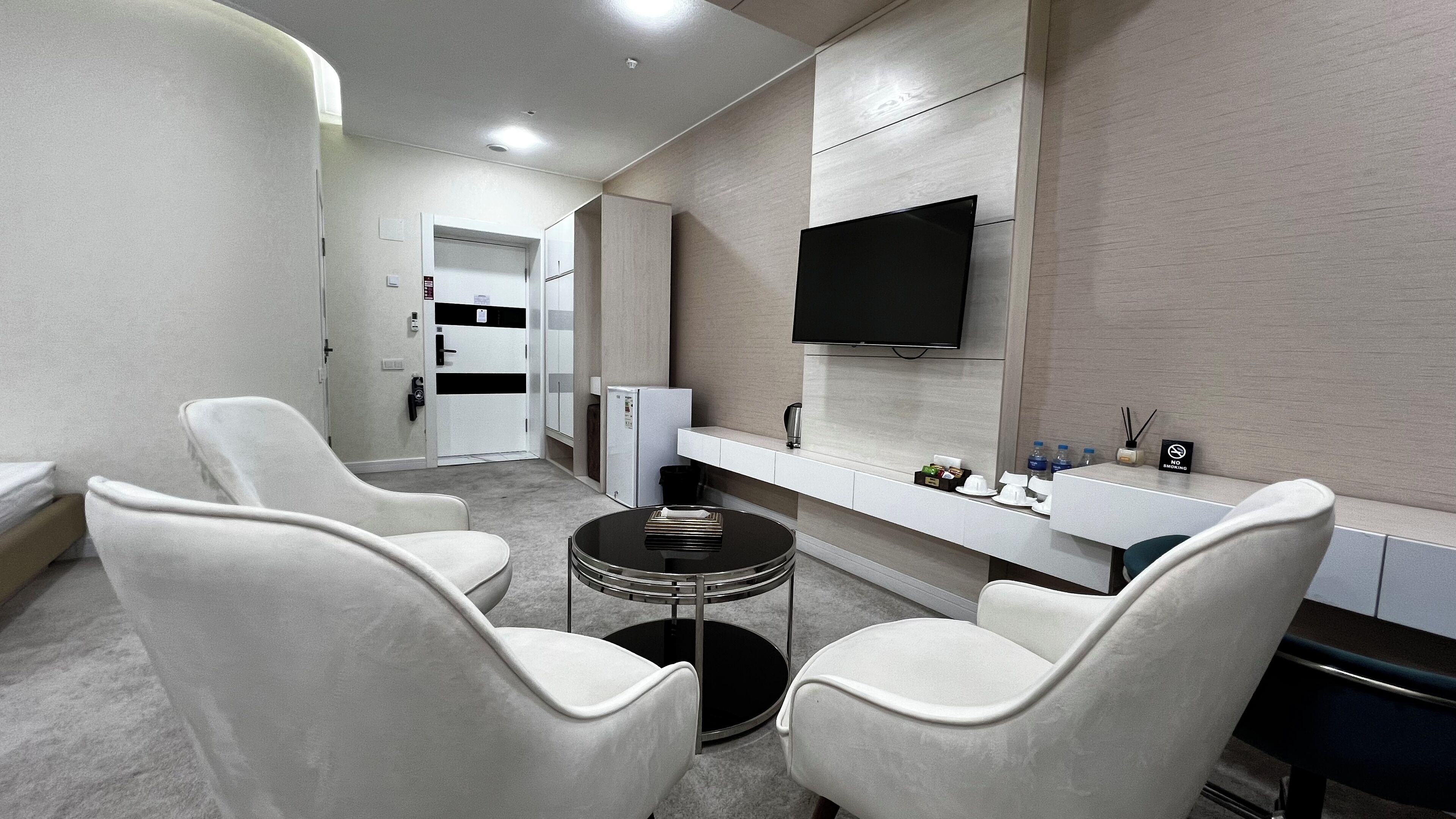 A modern hotel room with a flat-screen TV, a small round table, two white armchairs, a minibar, and a bathroom visible through an open door.