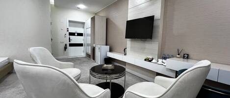 A modern hotel room with a flat-screen TV, a small round table, two white armchairs, a minibar, and a bathroom visible through an open door.