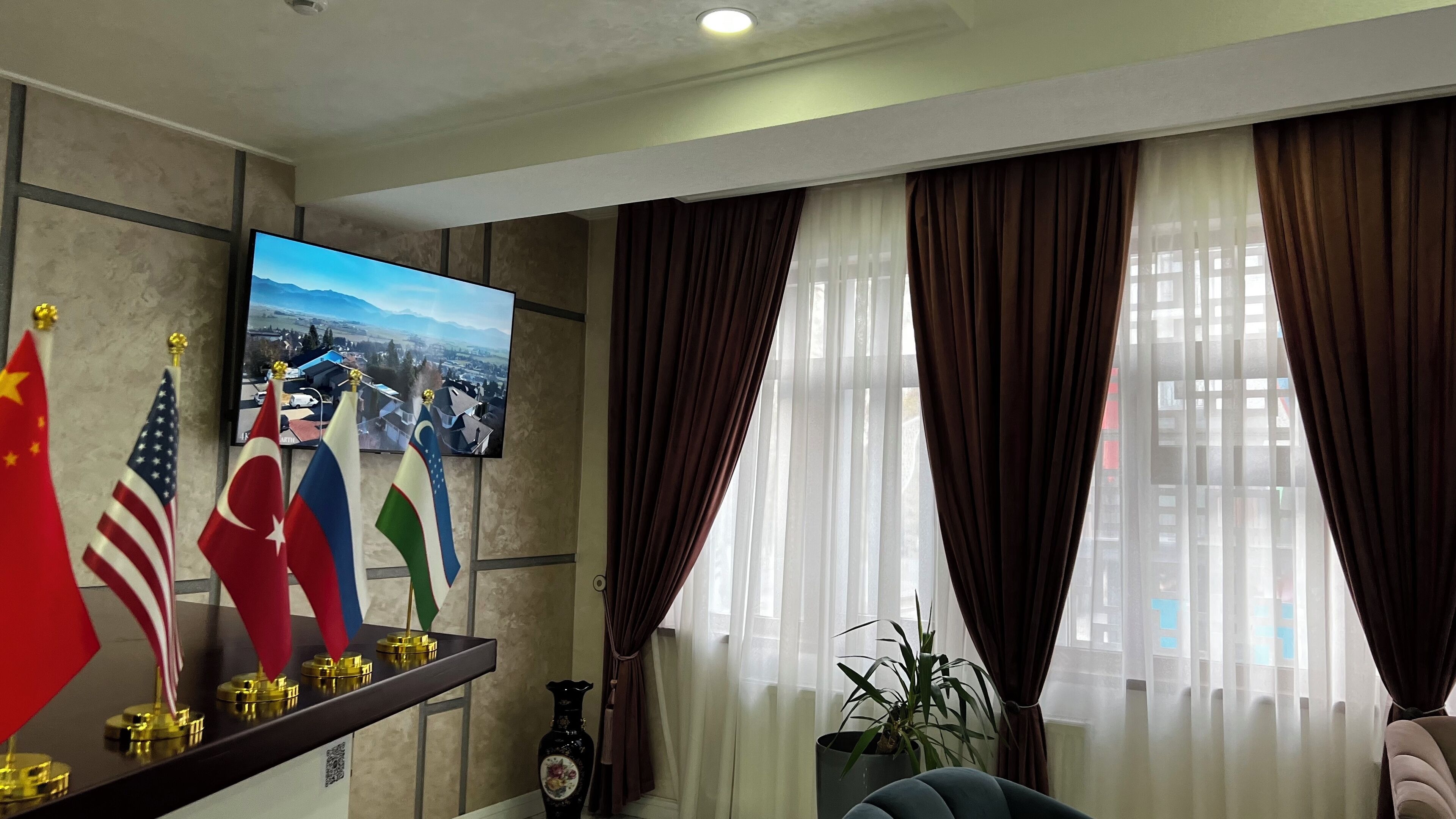 Una sala de estar con un televisor de pantalla plana mostrando una vista panorámica, un estante con banderas internacionales y un ventanal con cortinas.