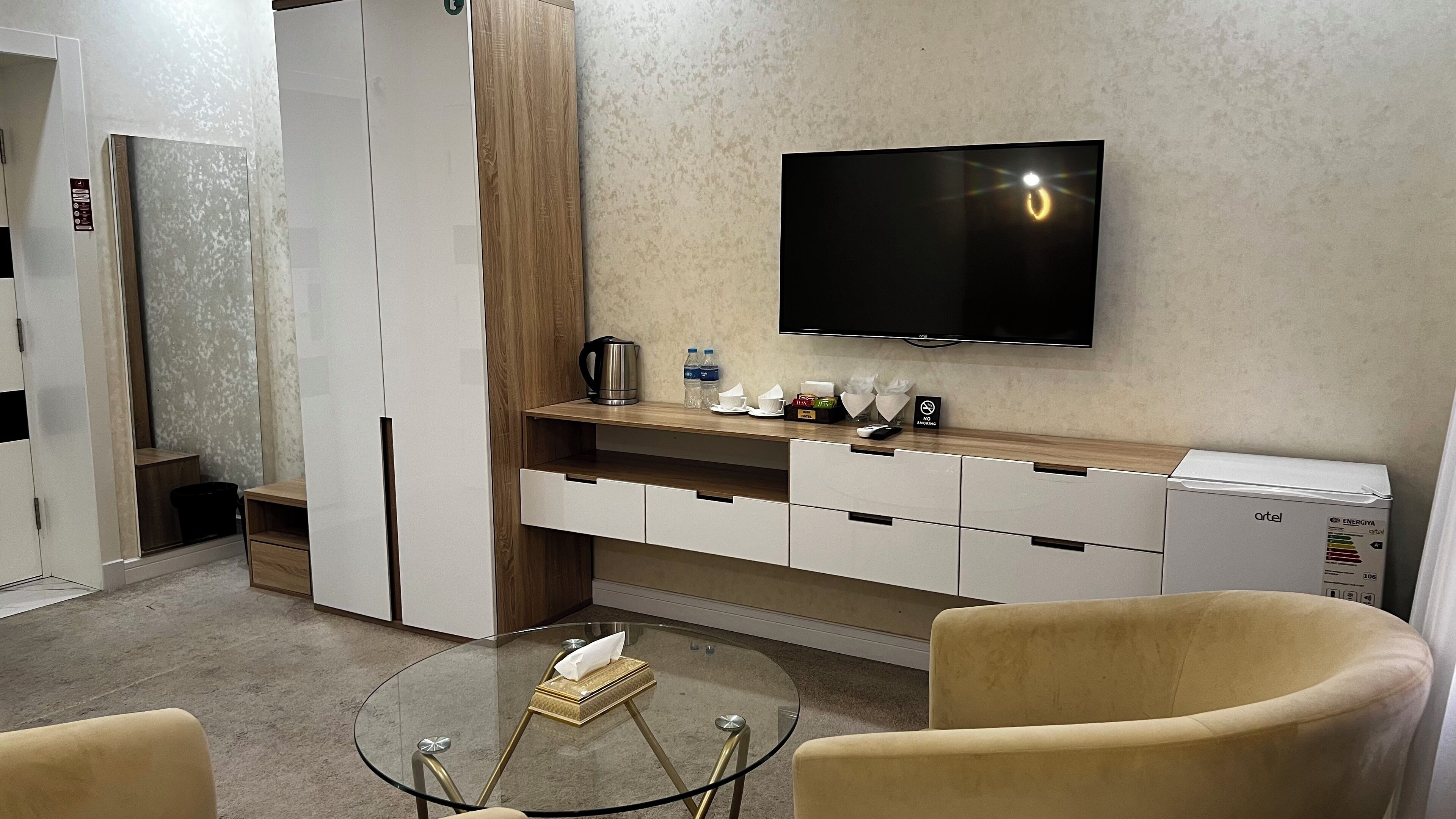 A modern hotel room with a flat-screen TV, a minibar, a glass coffee table, and a sofa.