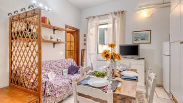 A cozy living room with a floral sofa, a dining table set for two, a television, and a kitchen area with a refrigerator.
