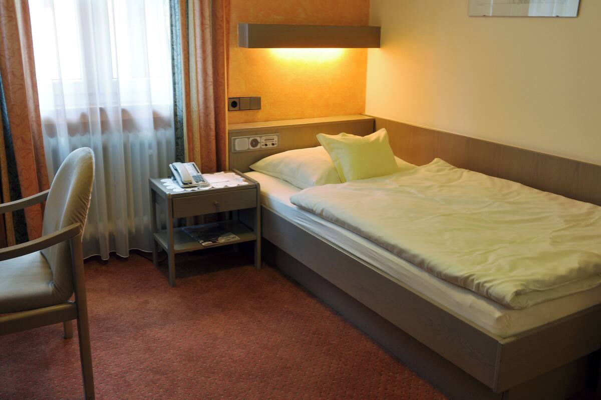Une chambre d’hôtel avec un lit simple, une table de chevet avec une lampe, une chaise et une fenêtre avec des rideaux.