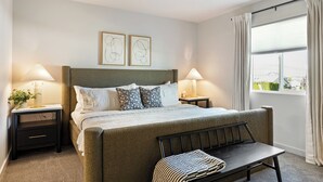 A bedroom with a bed, two bedside tables, a bench, a window with curtains, and two wall art pieces.