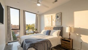 Une chambre à coucher avec un grand lit, une table de chevet, une lampe et une vue sur la ville grâce à de grandes fenêtres.