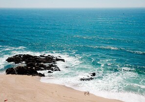 Una playa con agua azul cristalina, afloramientos rocosos y algunas personas caminando.