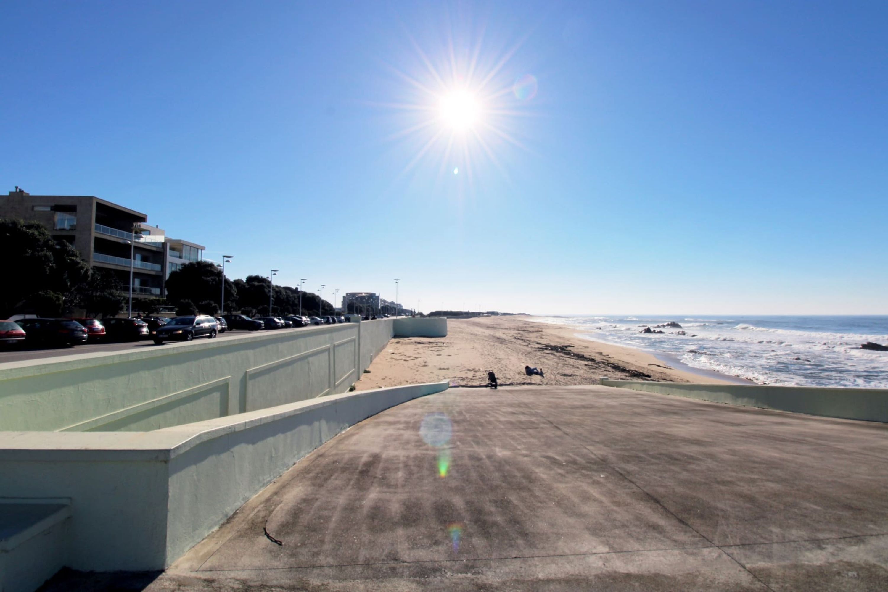 A beach with a concrete walkway, cars parked along the side, and a building in the background.