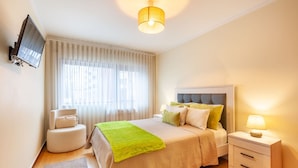 A bedroom with a bed, bedside table, TV, and a window with curtains. - Apartamento Hibiscos - Minho s Guest (Braga)