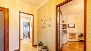 A hallway with wooden flooring, a welcome sign, potted plants, a mirror, and a white cabinet with a shelf. - Apartamento Hibiscos - Minho s Guest (Braga)