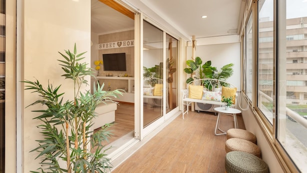 A balcony with a view of a cityscape, a sliding glass door leading to a living area with a sofa and a TV, and a small table with a plant.