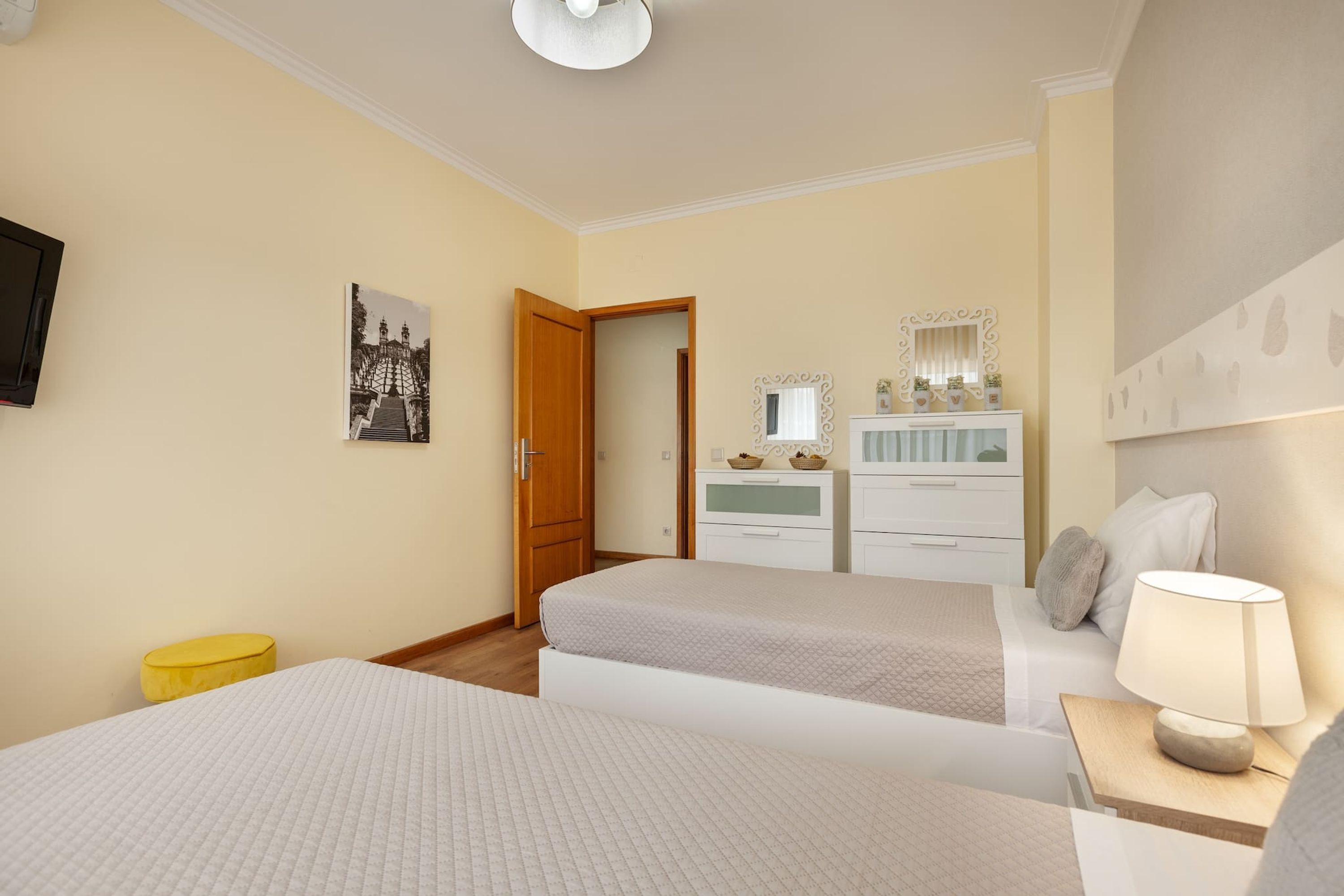 Une chambre d’hôtel avec deux lits, une porte en bois, une commode blanche avec un miroir, une petite table avec une lampe et un pouf jaune.