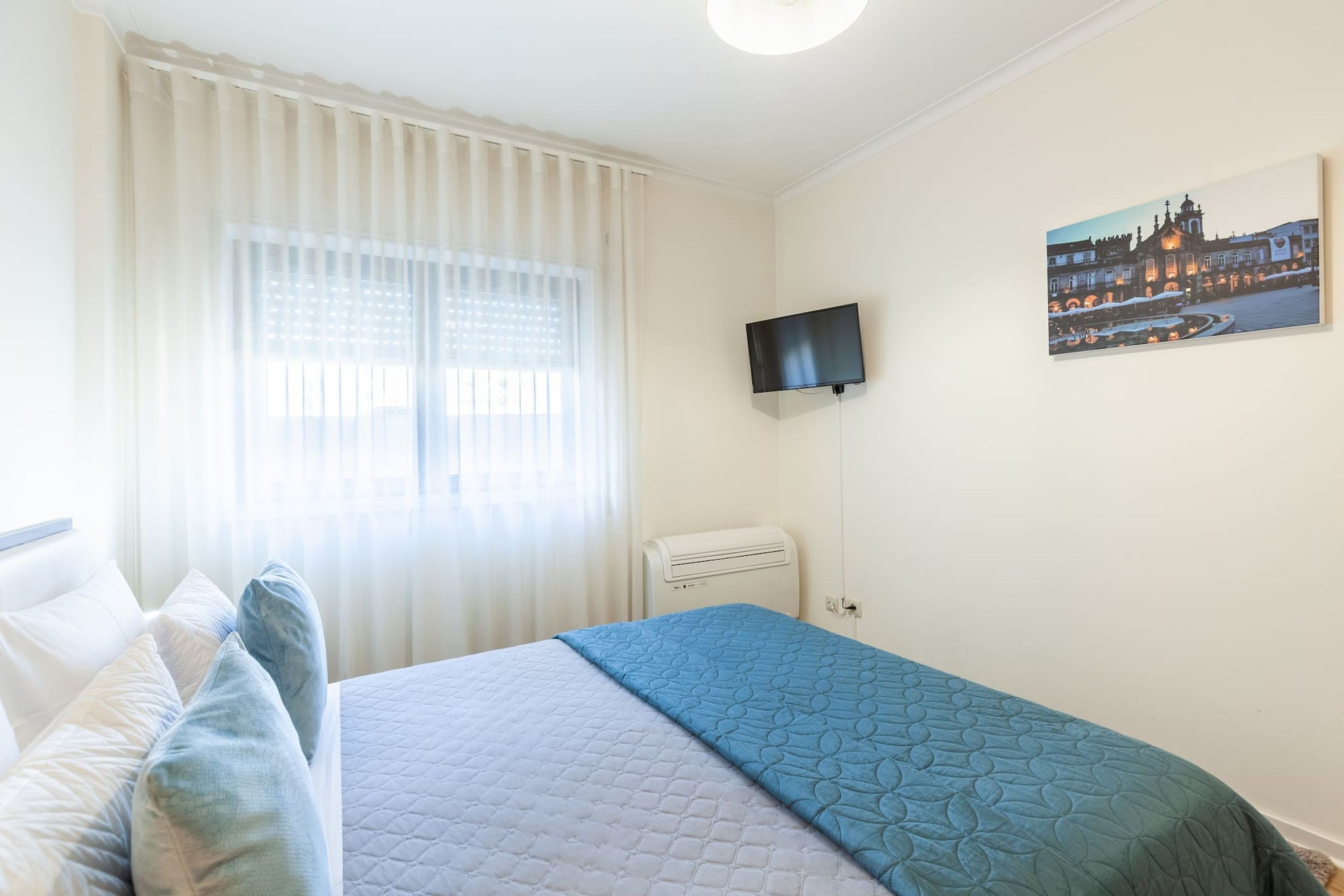 A hotel room with a bed, a TV mounted on the wall, and a painting of a cityscape.