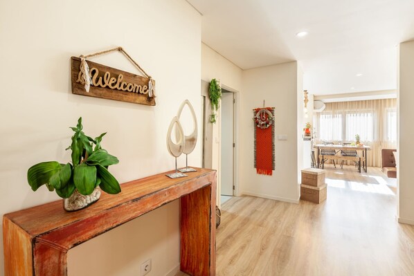 A modern living room with a wooden console table, a welcome sign, a potted plant, and a dining area in the background. - Apartamento L rio - Minho s Guest (Braga)