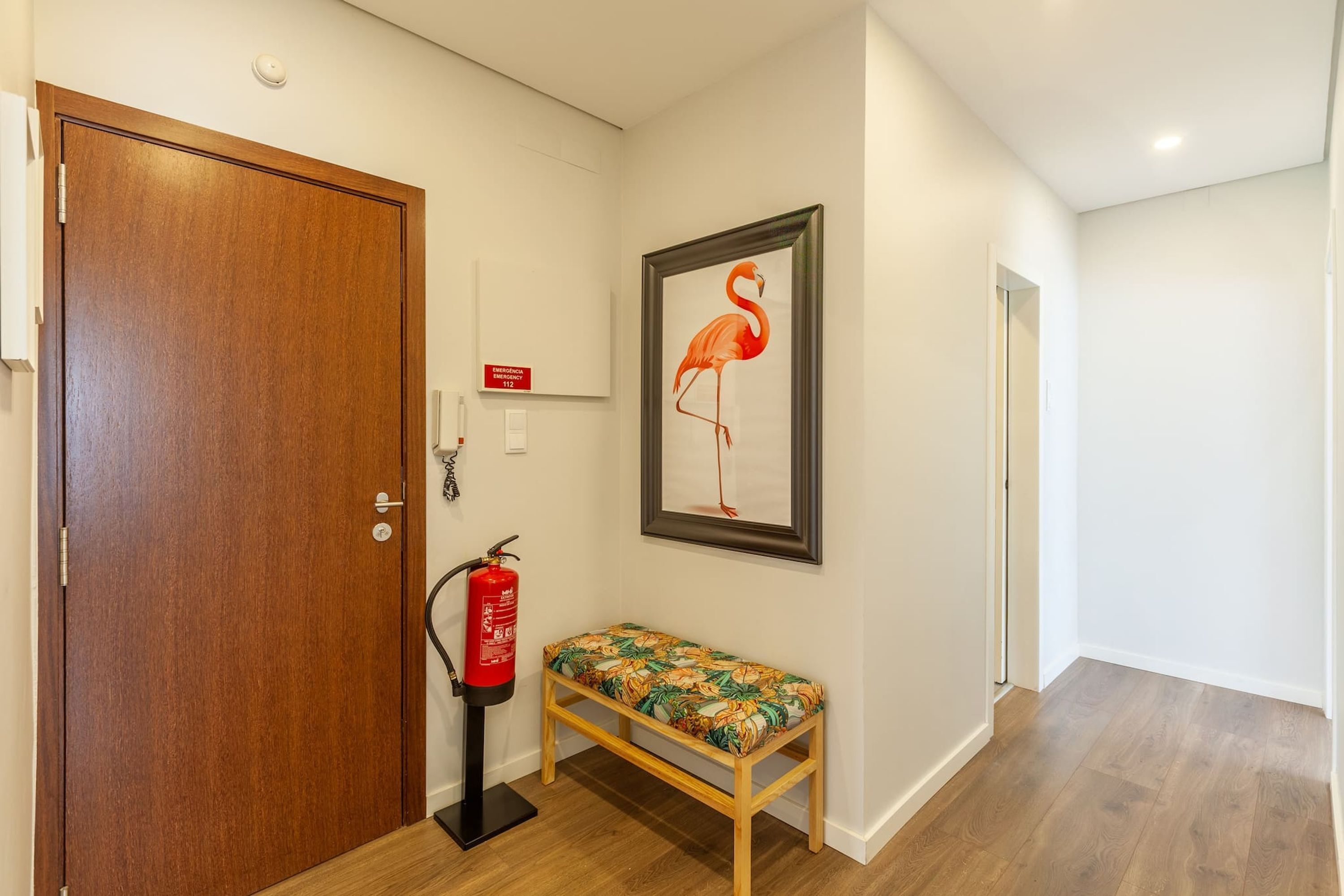 Un pasillo con una puerta de madera, un extintor, un banco y un cuadro enmarcado de un flamenco en la pared.