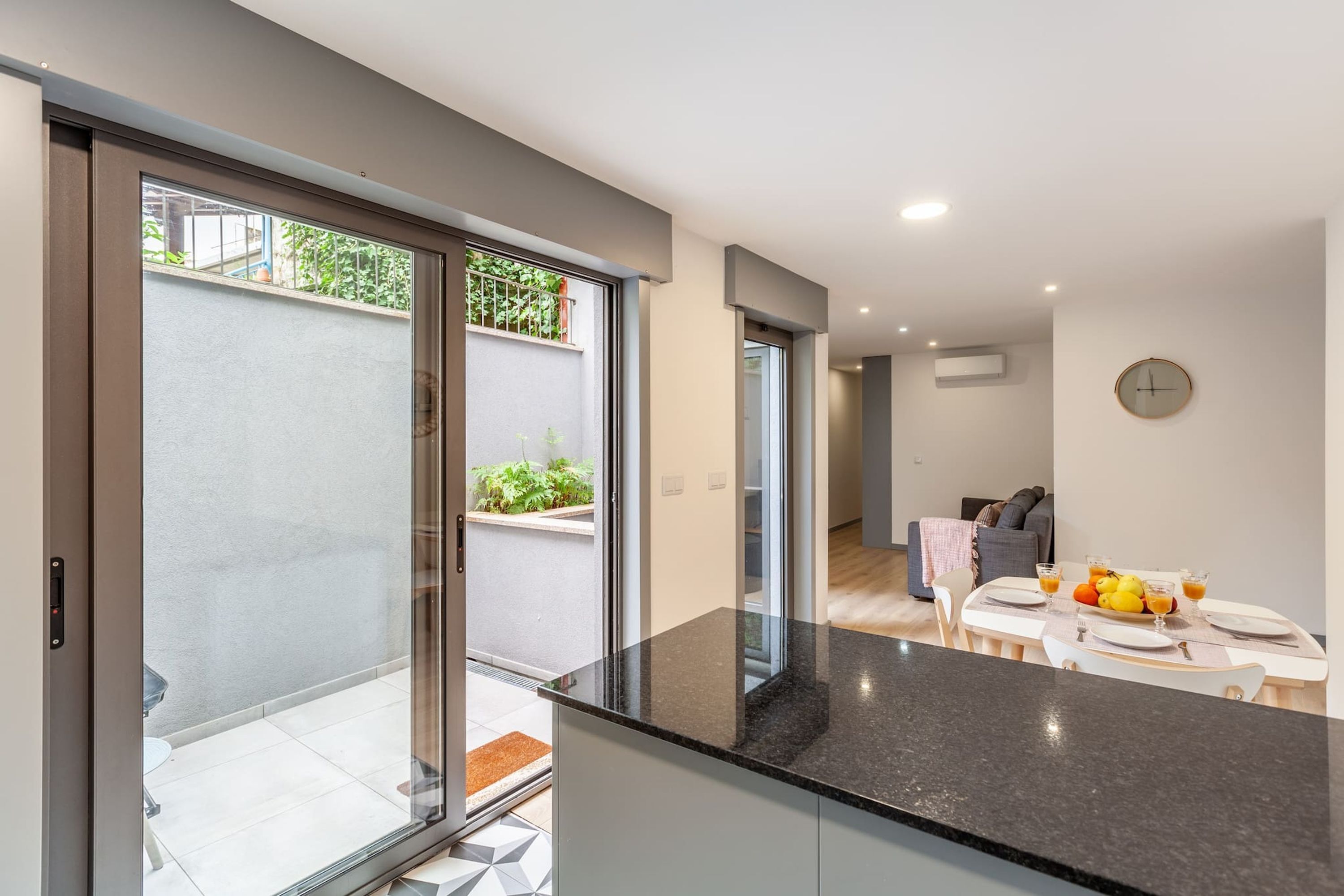 A modern kitchen with a black countertop, a dining area with a table set for two, and a sliding glass door leading to an outdoor area with plants.