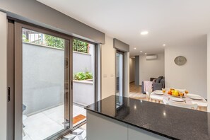 Une cuisine moderne avec un plan de travail noir, un coin repas avec une table dressée pour deux personnes, et une porte-fenêtre coulissante donnant sur un espace extérieur agrémenté de plantes.