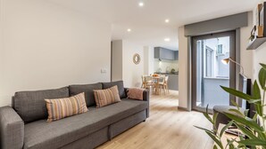 Un salon moderne avec un canapé gris, un coin repas et une cuisine visible à travers une porte ouverte.