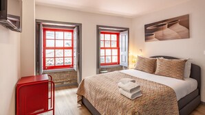 Une chambre à coucher avec un lit, deux fenêtres aux cadres rouges, une chaise rouge et un tableau accroché au mur.