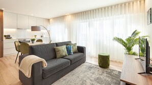 Un soggiorno moderno con un divano grigio, un poggiapiedi rotondo, un tavolino da caffè in legno e una TV. C'è un angolo cottura con mobili bianchi e un tavolo da pranzo.