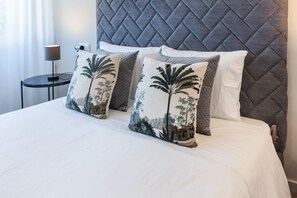 Un lit avec une tête de lit matelassée, deux oreillers ornés de motifs de palmiers et une table de chevet avec une lampe.