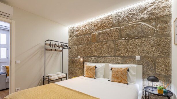 Un dormitorio con cabecera de pared de piedra, cama con manta estampada, mesita de noche con lámpara y perchero con ropa colgada.