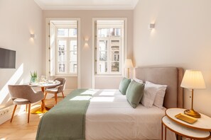 A hotel room with a bed, a bedside table with a lamp, a chair, a small table with a plant, and a window with a view of buildings. - Living Valverde Studio Verde - Minho s Guest (Braga)