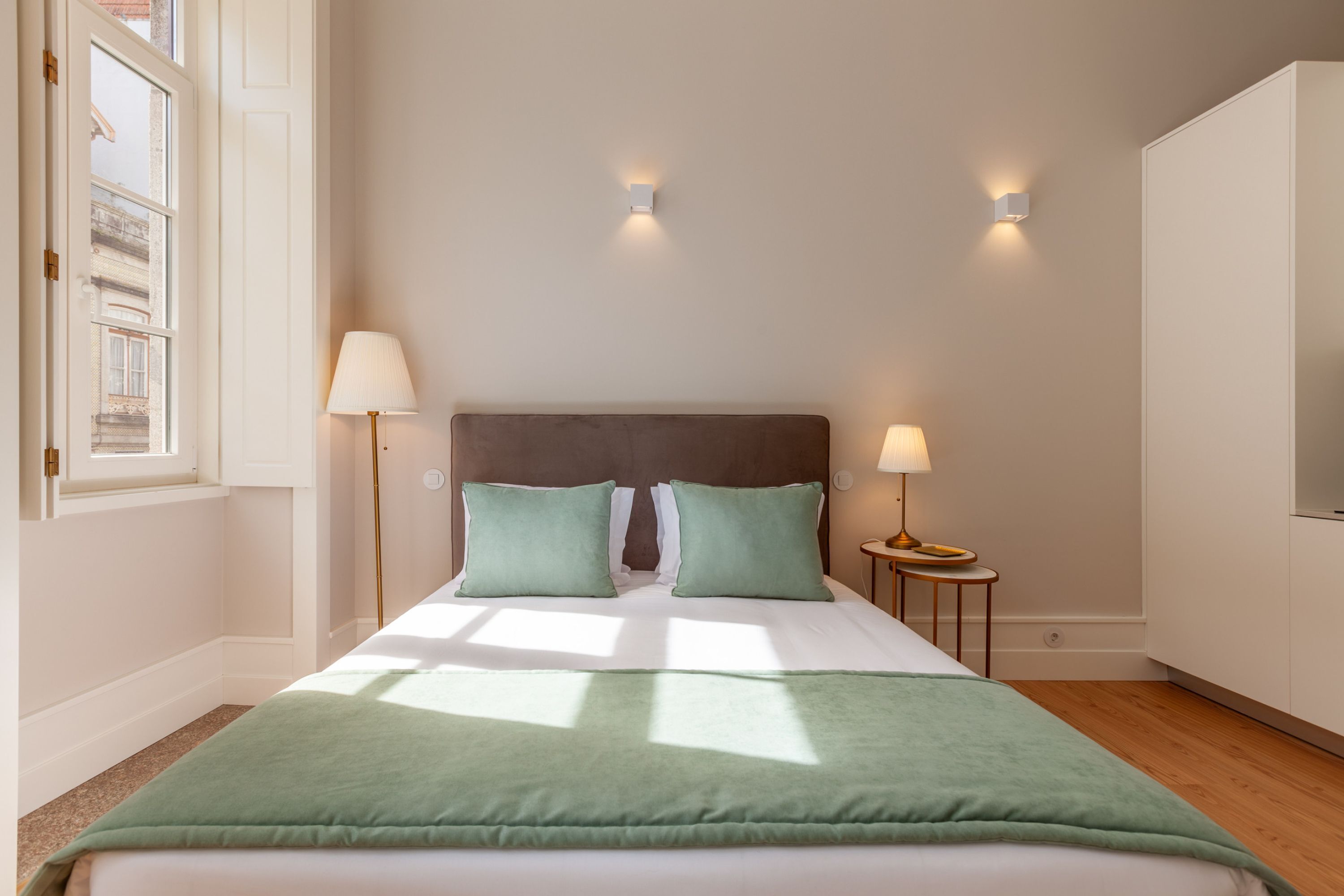 Una cama bien tendida con una manta verde, dos lámparas de noche y una mesita con lámpara.