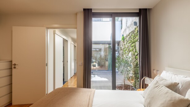 Ein Schlafzimmer mit einem großen Bett, einer Schiebetür zu einem Balkon mit Blick auf eine Steinmauer und Grünpflanzen.