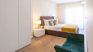 Hotellihuone, jossa on sänky, yöpöytä, vihreä tuoli ja suuri ikkuna, jossa on verhot.