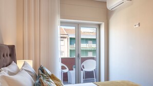 Hotellihuone, jossa on sänky, ikkuna verhoilla, ilmastointilaite sekä parveke, jossa on tuoleja ja pöytä.