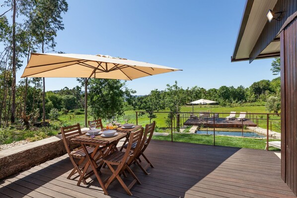 A wooden deck with a table and chairs, an umbrella, and a view of a pool and garden.