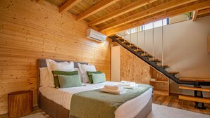 Une chambre à coucher aux murs lambrissés, avec un lit, des oreillers, un plaid vert, un banc en bois et un escalier.