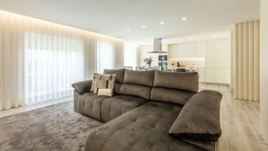 Una sala de estar moderna con un sofá seccional grande, una cocina al fondo y amplias ventanas con cortinas de gasa.