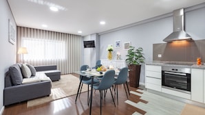 A modern kitchen with a built-in oven, a dining area with a glass table and chairs, and a grey sofa with a lamp. - Libert Lodge Apartment - Minho s Guest (Braga)