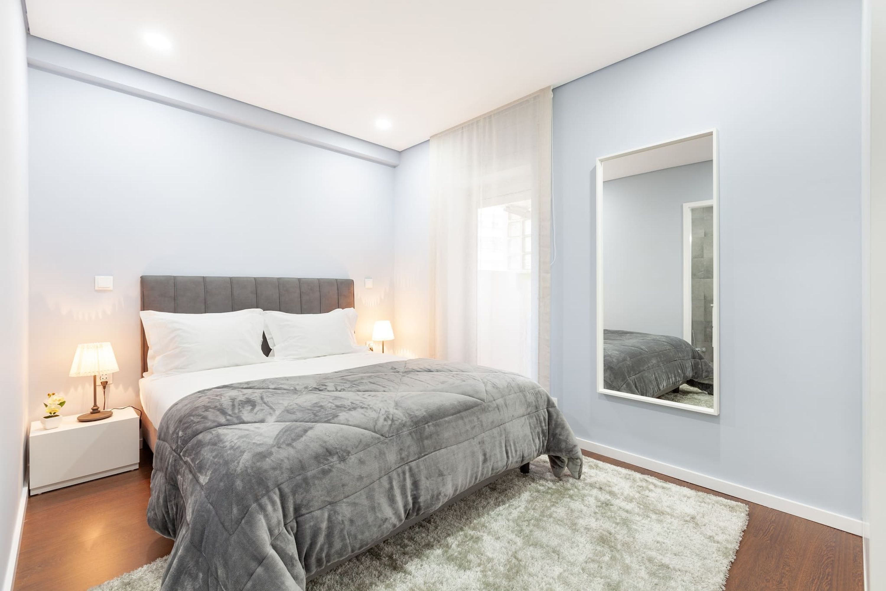 A modern bedroom with a grey bed, white nightstands, and a large mirror.