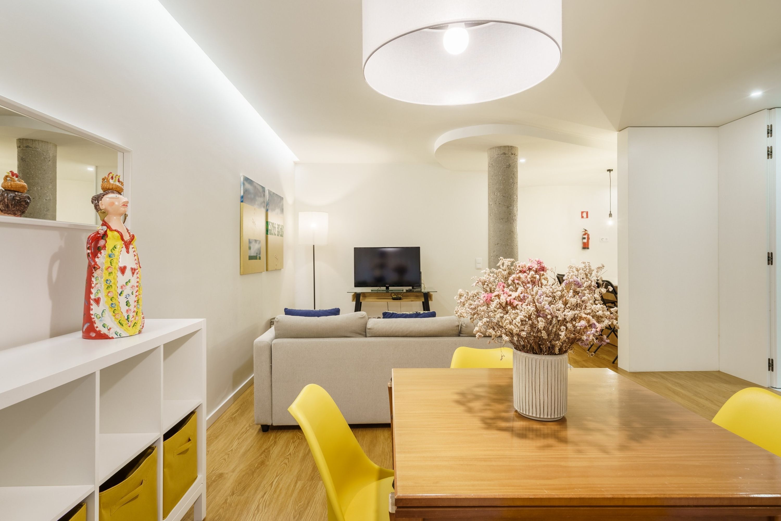 Una sala de estar moderna con una mesa de comedor de madera, sillas amarillas, un sofá, un televisor y objetos decorativos, incluyendo una estatua y un jarrón con flores.