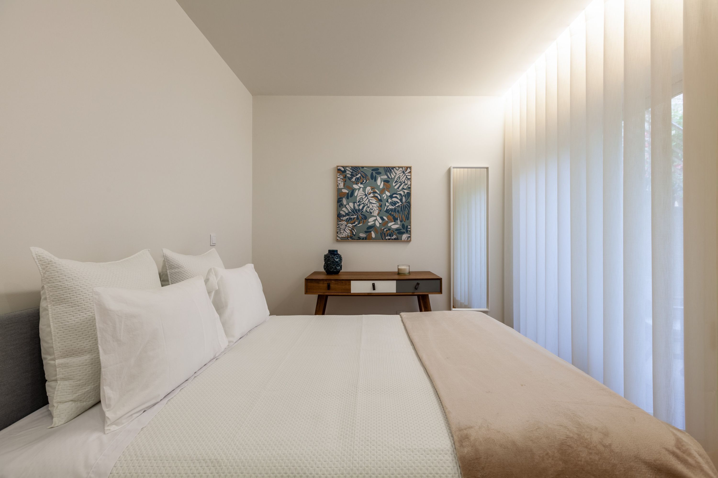 A bedroom with a bed, pillows, a bedside table with a vase, a mirror, and a window with curtains.