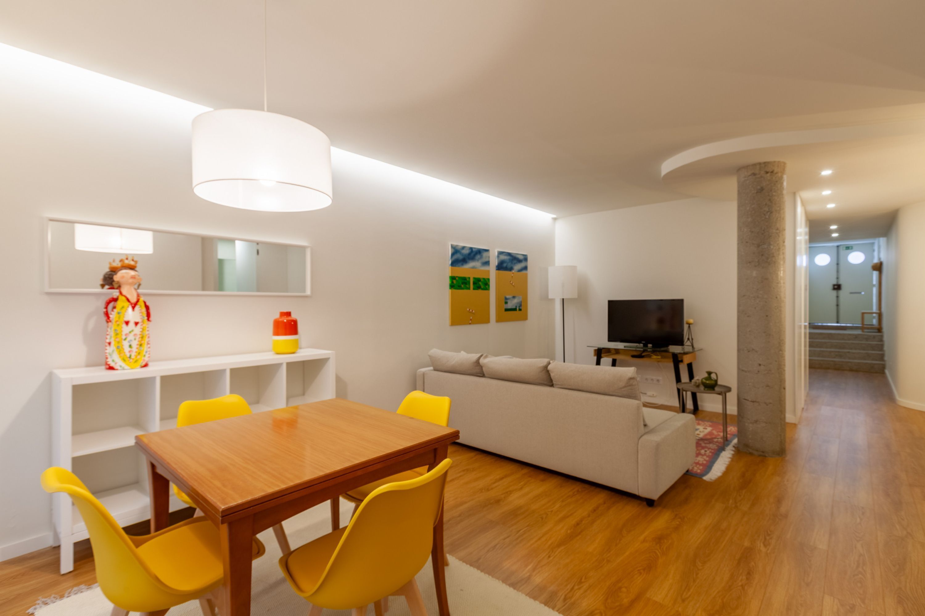 Un salon moderne avec une table à manger en bois, des chaises jaunes, un canapé beige, un téléviseur et un grand luminaire suspendu blanc.