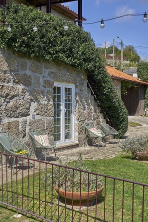 A stone house with a white door, two wicker chairs with cushions, a potted plant, and a metal fence. - Casa do Xico - Minho s Guest (Guimarães)