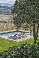 Una piscina con sillones de descanso, rodeada por un jardín cercado y un árbol de naranjos.