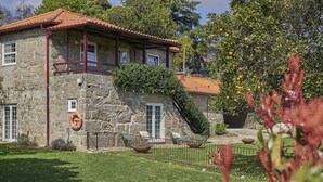 A stone building with a red-tiled roof, a balcony, and a grassy yard with a fence. - Casa do Xico - Minho s Guest (Guimarães)