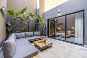 Un espacio al aire libre moderno con un sofá gris, una mesa de centro de madera y plantas verdes grandes.
