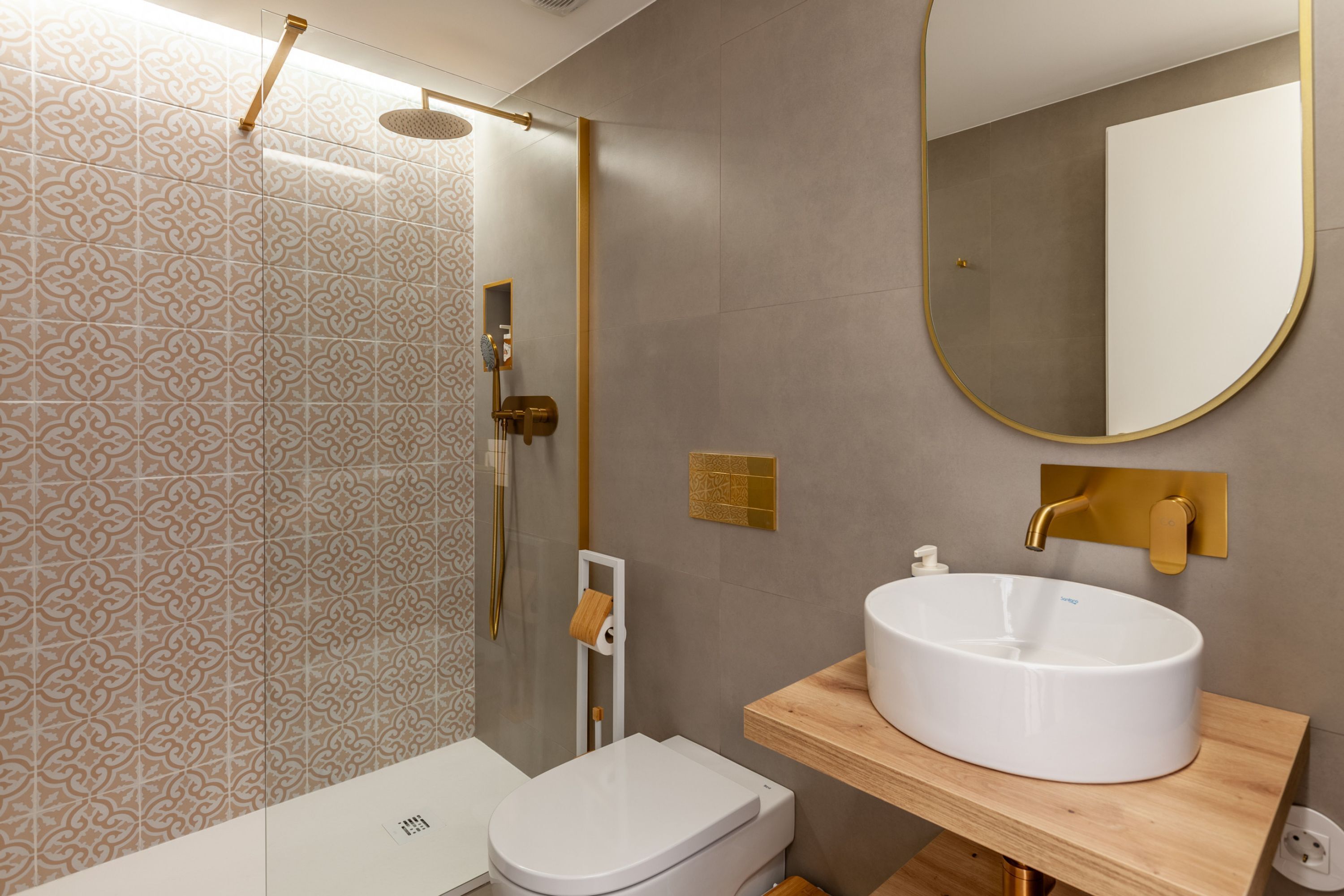 A modern bathroom with patterned tiles, a toilet, a sink with a gold faucet, and a mirror with a gold frame.