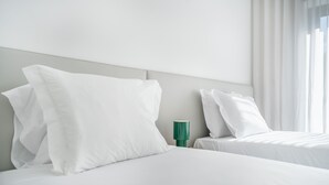 Camera d'albergo con due letti, biancheria da letto bianca, un comodino verde e tende leggere.