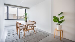 Un'area pranzo moderna con un tavolo e sedie in legno, una pianta in vaso e vista sugli edifici attraverso ampie finestre.