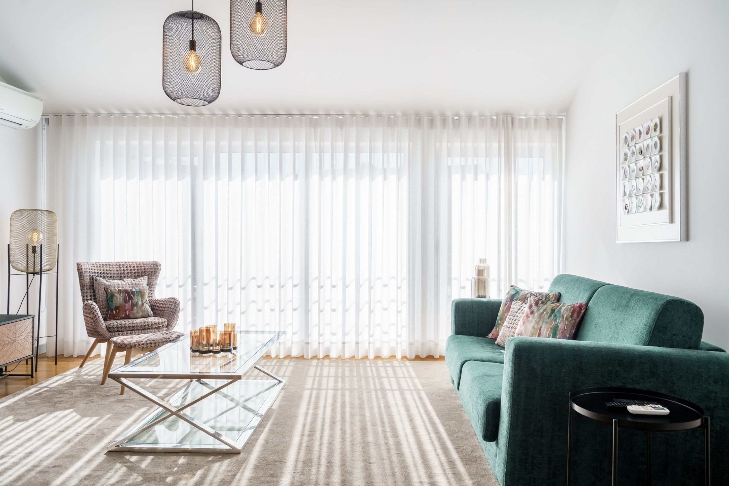 A modern living room with a green sofa, a glass coffee table, a patterned armchair, and a large window with sheer curtains.
