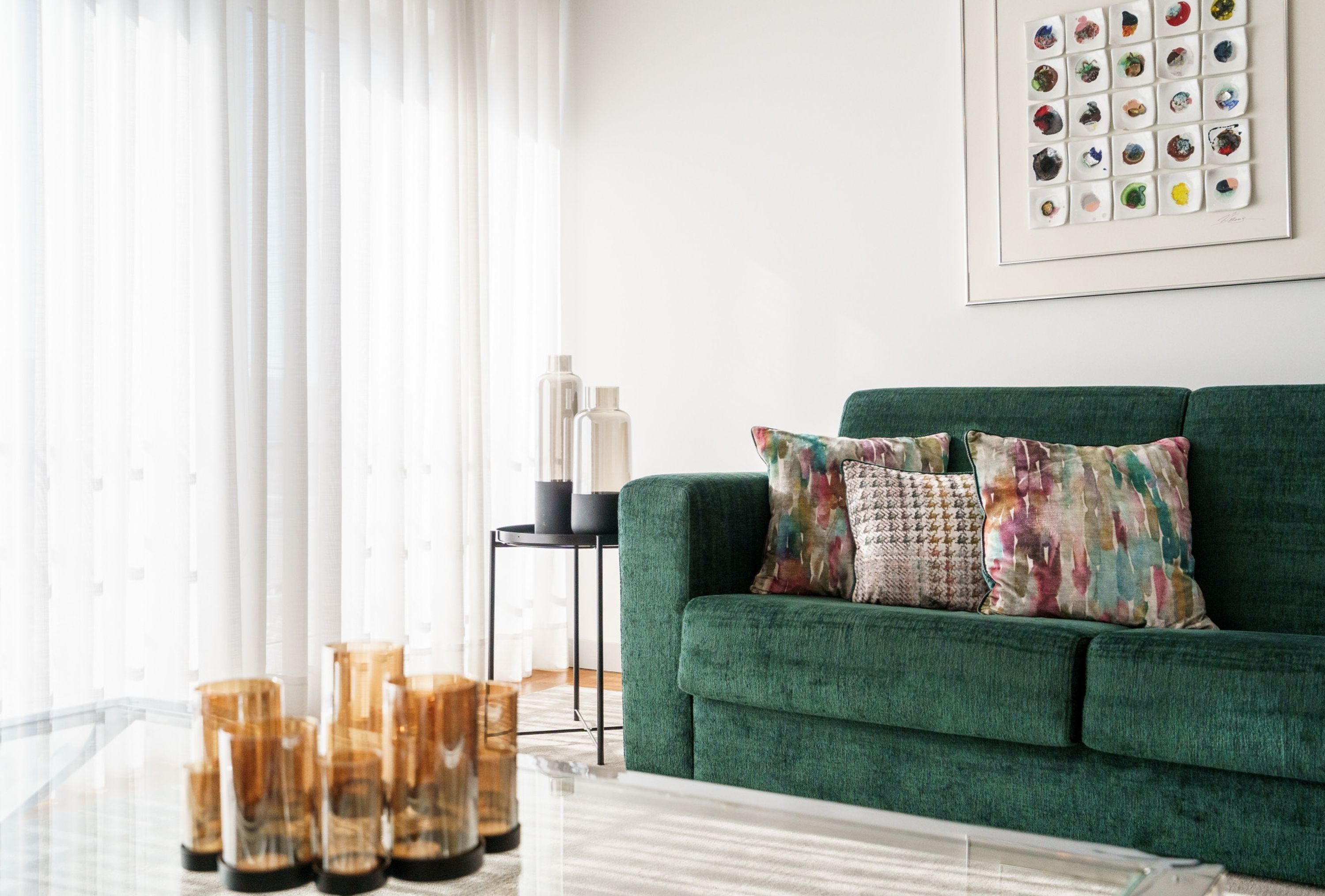 A modern living room with a green sofa, patterned cushions, a glass coffee table, and a framed artwork on the wall.